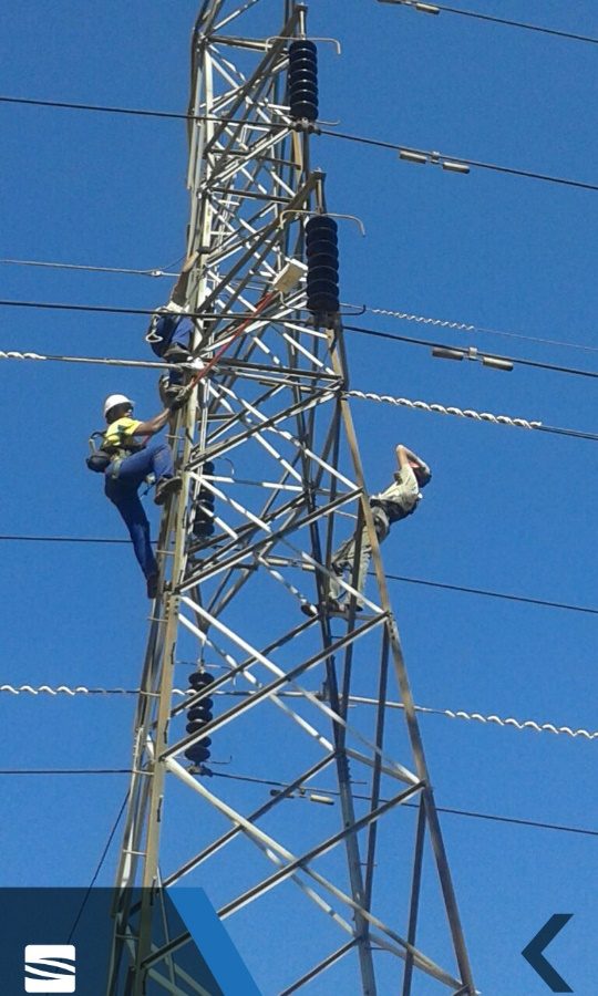 Two linesmen working on a steel transmission structure against a clear sky.