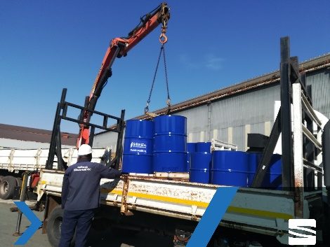 Steel drums of transformer oil being loaded by crane onto a flatbed truck.